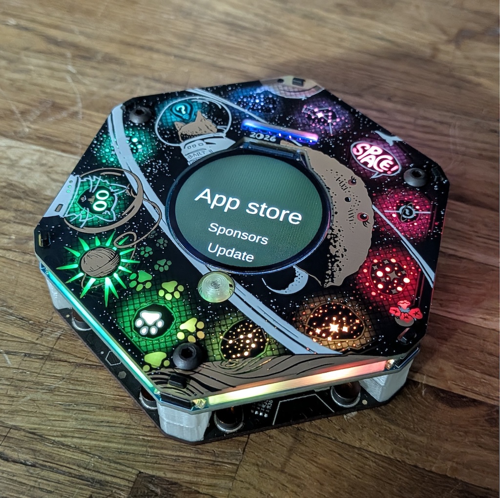 A photograph of the spaceagon. It is a hexagonal electronic badge covered in temporary artwork of space cats.